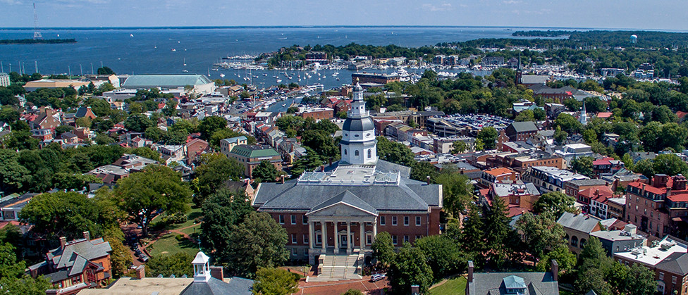 An aerial view of the City of Annapolis with the Maryland State House in the foreground, with buildings, trees, and the waterfront in the background.