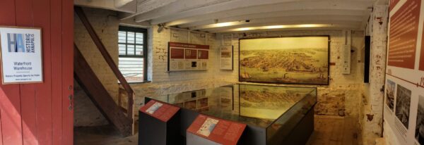 A red door opens into a room with whitewashed stone walls. On the far wall is a large painting of the Annapolis harbor as it looked in the 18th century. A large model of the harbor sits in the middle of the room enclosed in glass.