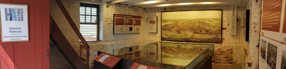 A red door opens into a room with whitewashed stone walls. On the far wall is a large painting of the Annapolis harbor as it looked in the 18th century. A large model of the harbor sits in the middle of the room enclosed in glass.