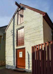 A brick building with no windows and a stone foundation that has been whitewashed. The building has three floors with three red, wooden panel solid doors, with a red wooden fence to the right of the building.