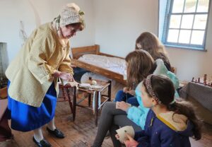 An educator in period dress shares sewing items with children