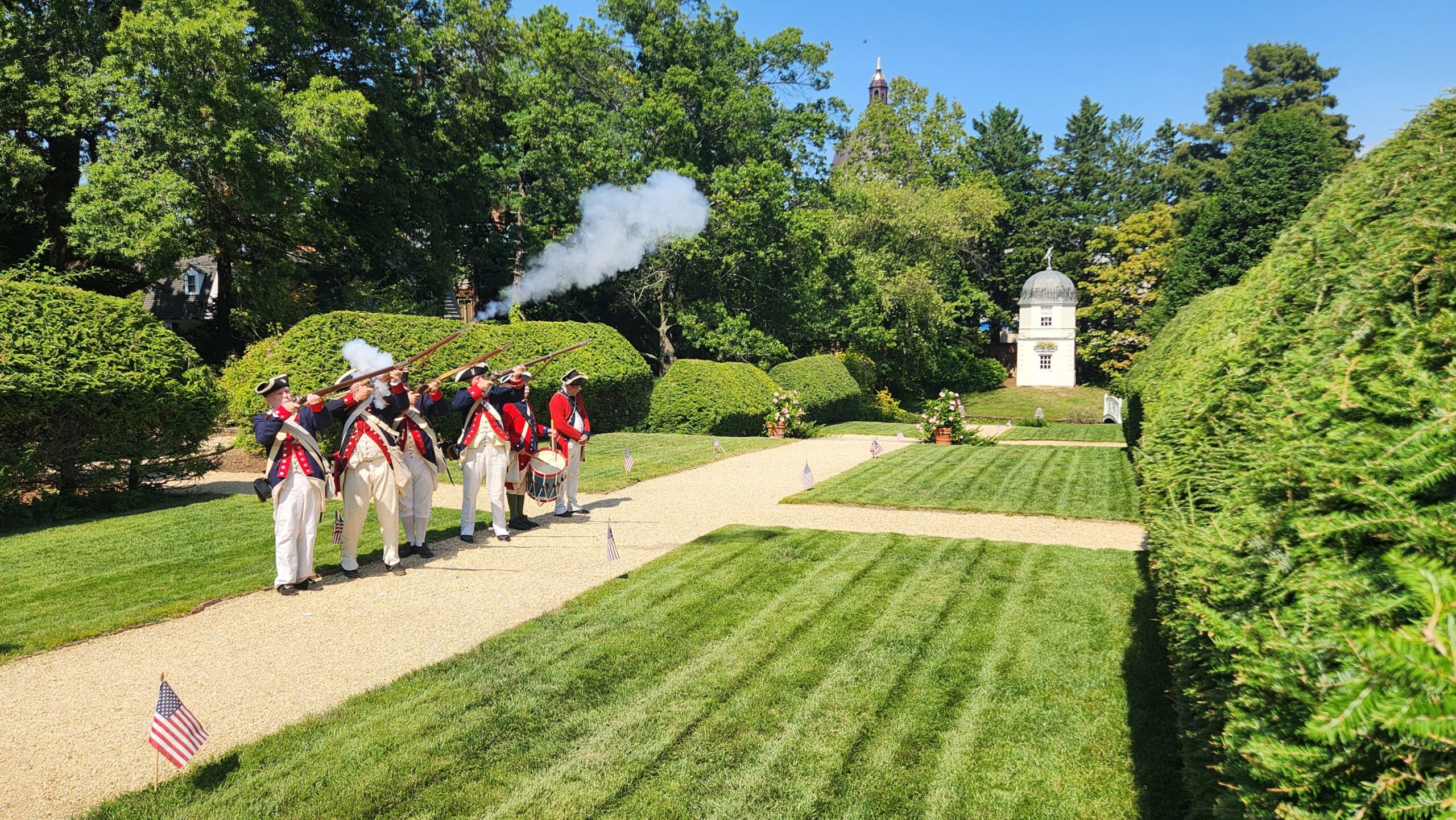 Four historic reenactors performing a musket salute in a historic garden.