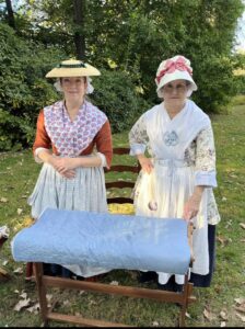 Two women in colonial period dress smile for the camera in an outdoor setting